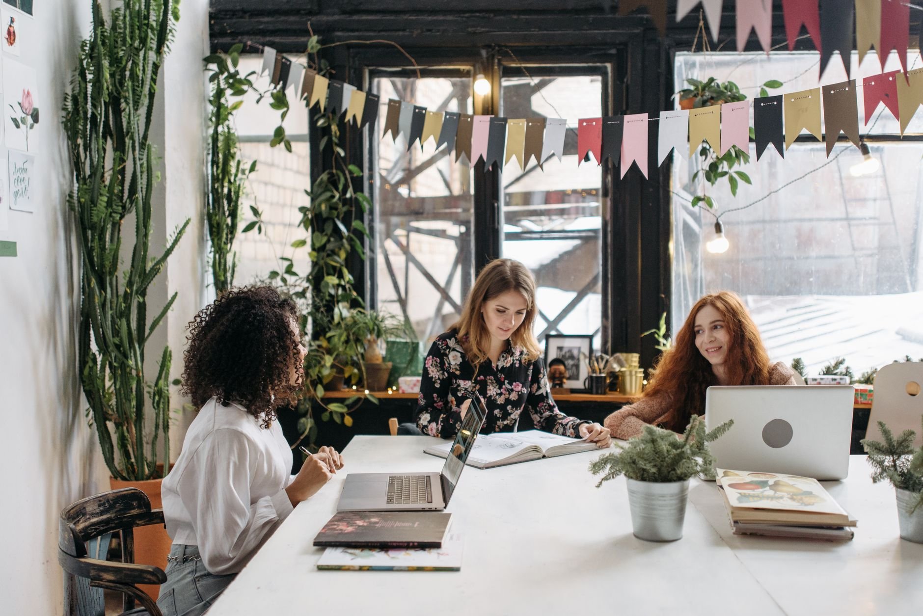 Three women working collaboratively in a stylish, plant-filled workspace featuring laptops and natural light.